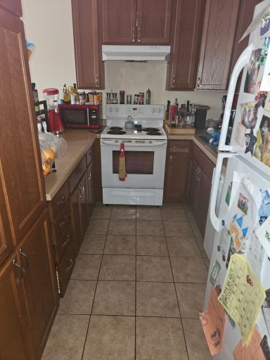 Narrow kitchen view with wooden cabinets, stove, refrigerator, and tiled floor.